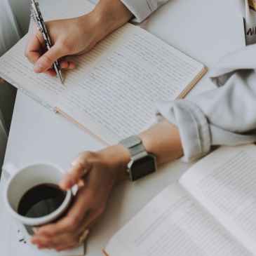 person writing on notebook while holding coffee mug